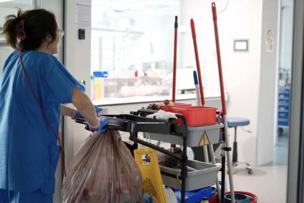 Housekeeper wearing blue uniform and gloves pushing cleaning trolley