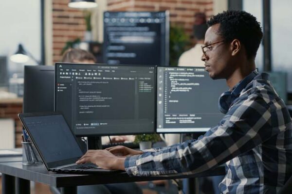 Portrait of african american developer using laptop to write code sitting at desk with multiple screens parsing algorithm in software agency. Coder working on user interface using portable computer.